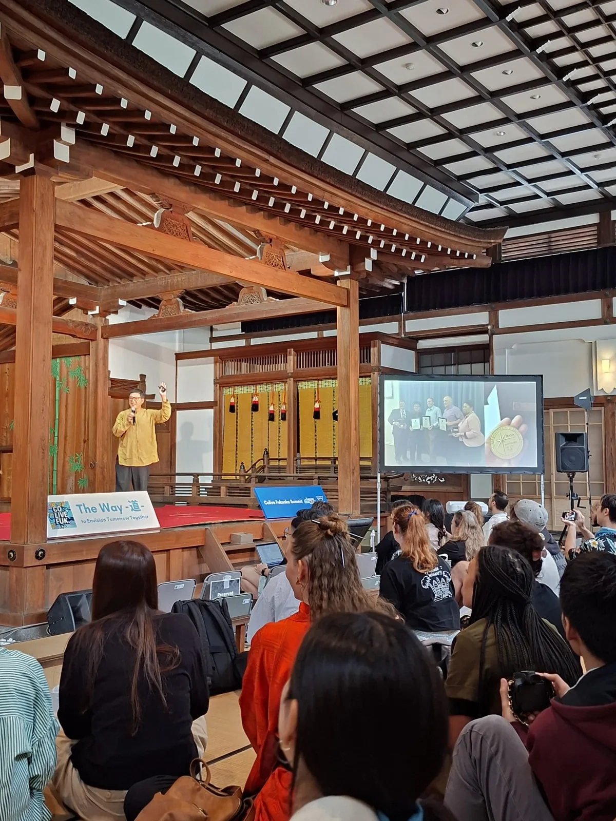 A speaker on a traditional wooden stage addressing the Colive Fukuoka Summit audience seated on tatami.