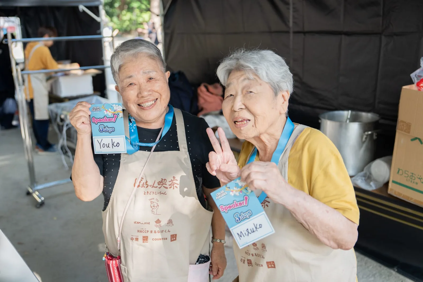 Two elderly Japanese women holding Colive Fukuoka speaker badges and smiling at the camera.